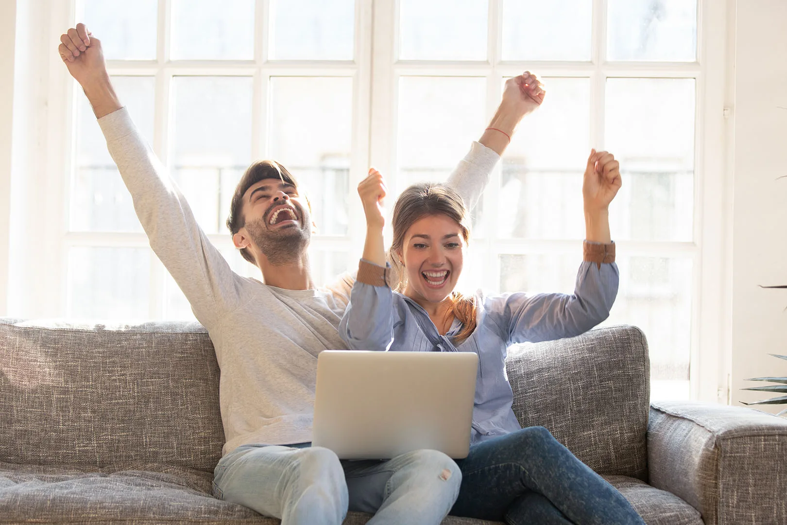 A man and woman sit on a couch, arms raised in excitement, celebrating a quick cast cash purchase completed effortlessly on their laptop.
