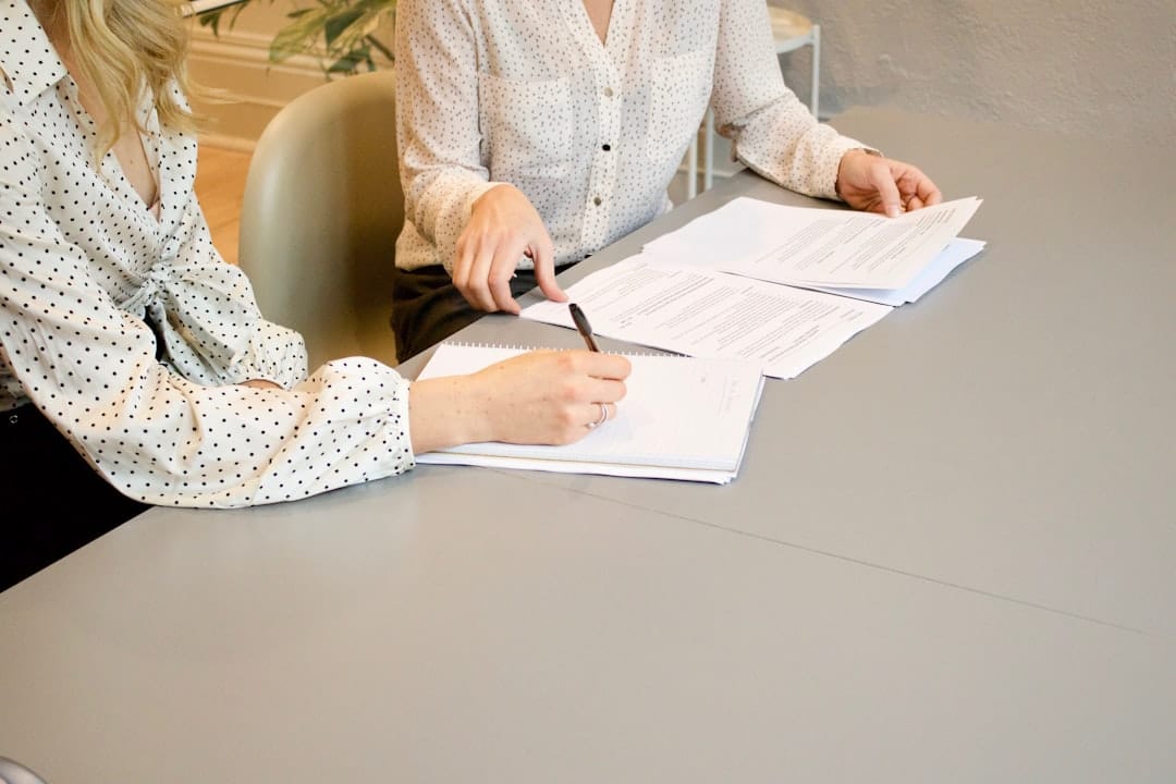 Two people sit at a table reviewing documents. One points at a page, while the other holds a pen, their dotted blouses adding a touch of charm. They discuss the cash purchase details to ensure fast transaction processing for their client.