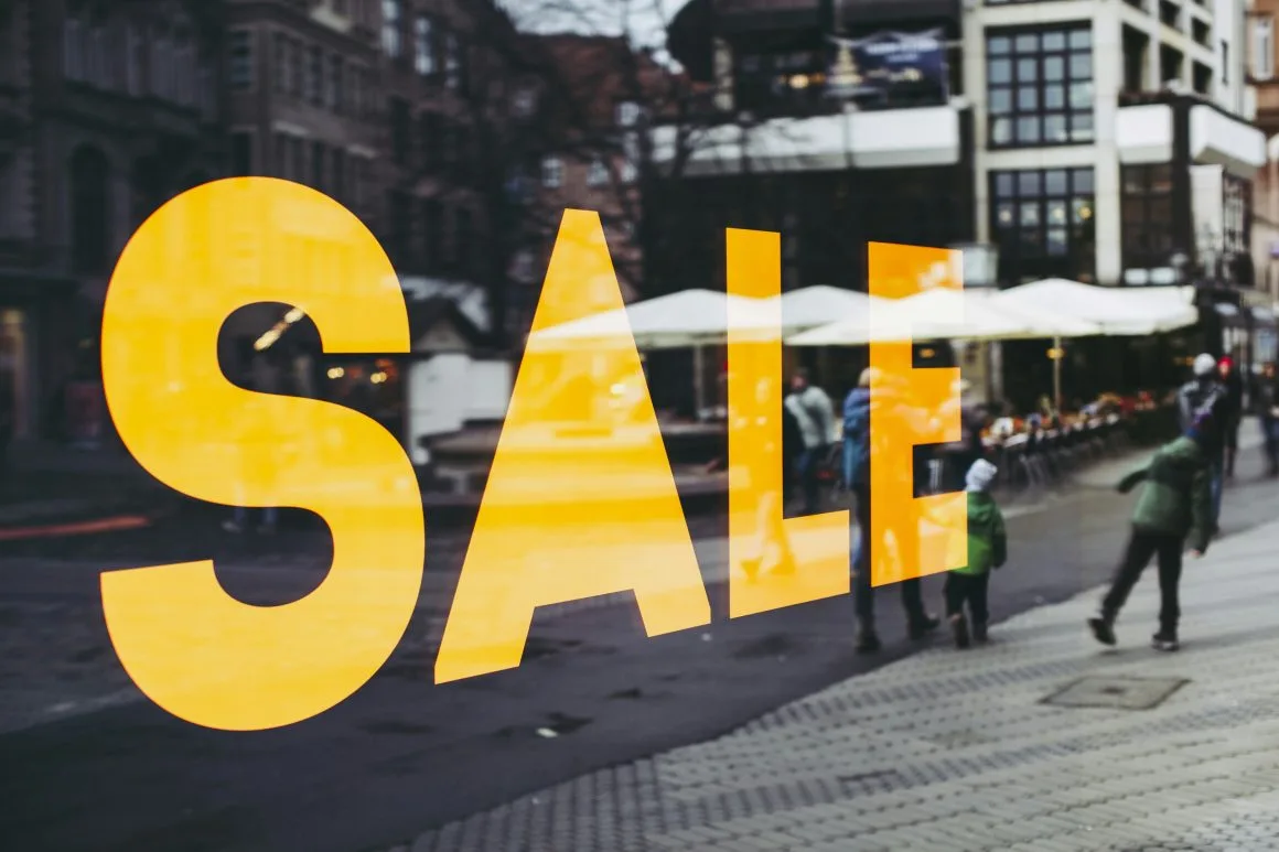 A large yellow "SALE" sign dominates the shop window, reflecting a bustling street scene where pedestrians and outdoor seating abound. Ideal for quick transactions or fast cash purchases, this lively tableau captures the buzz of urban life.