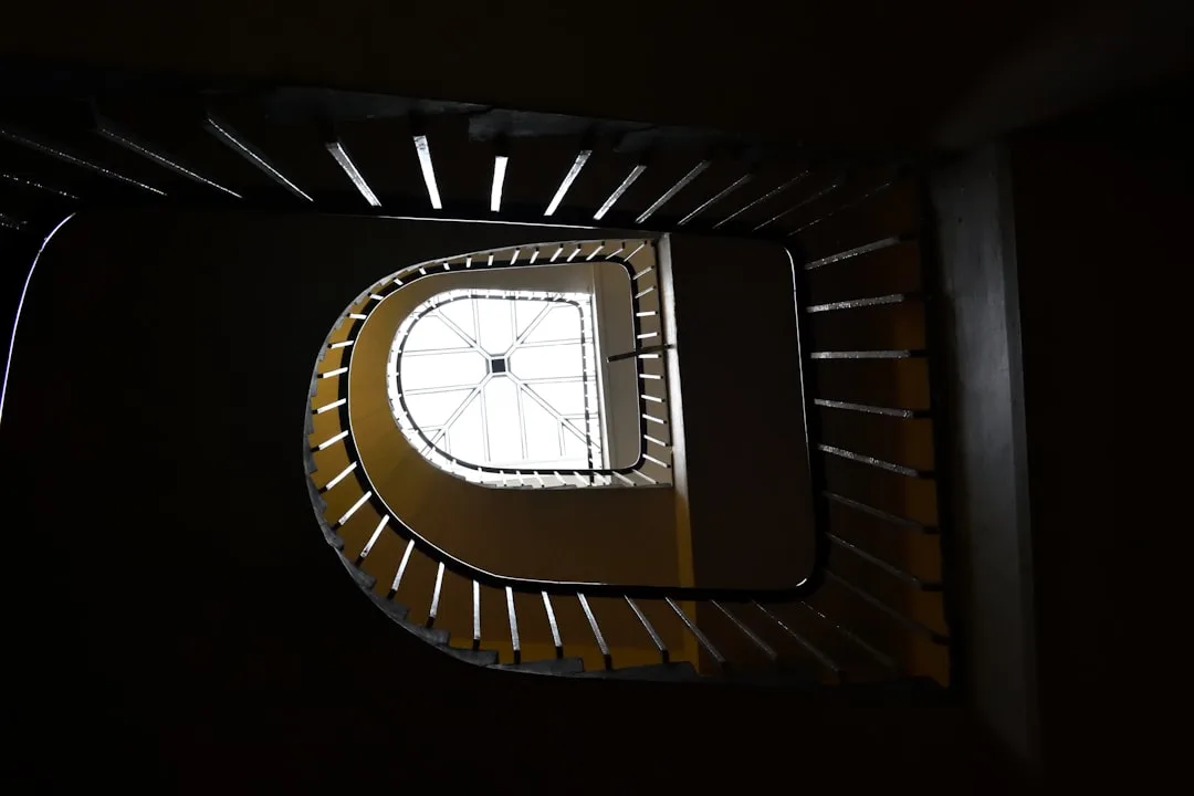 View looking up a spiral staircase with a window at the top, casting light and shadow on the steps and walls, reminiscent of a quick cast of light.