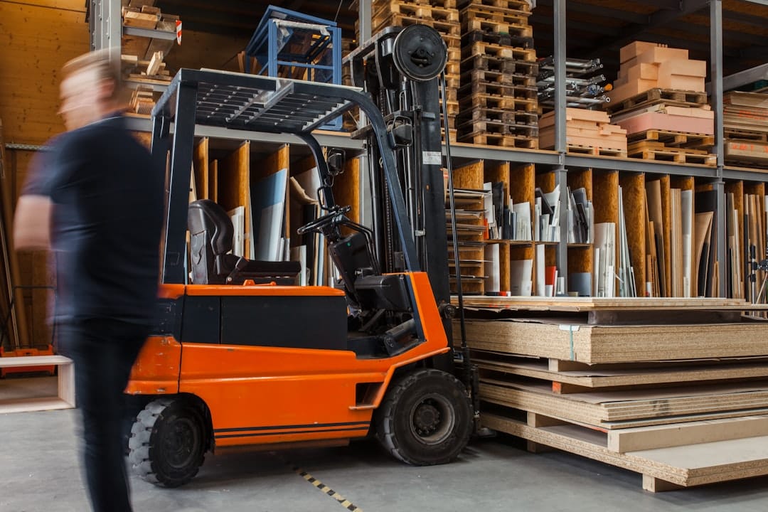 A person walks past an orange forklift in a lumber warehouse, where stacks of wood and various materials are stored on shelves, ready for fast transaction.