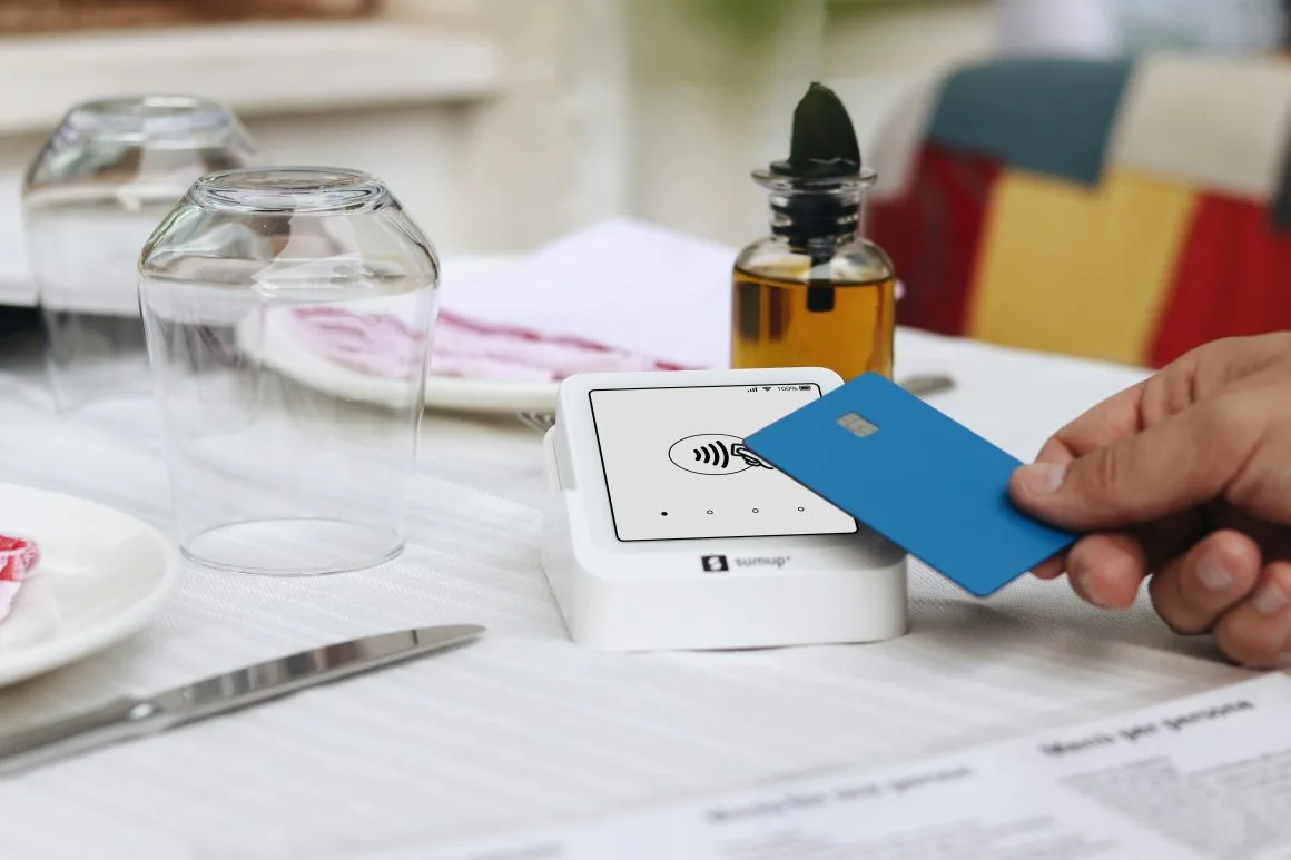 A person swiftly hovers a blue contactless card above a white card reader on a table adorned with glassware and a menu, enabling a fast transaction.